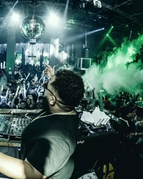 DJ with hands raised at the decks facing a festival crowd under green lights
